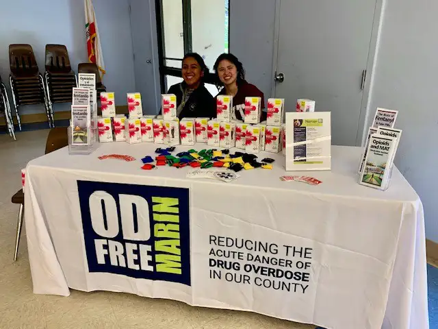 Two women sitting behind an OD Free Marin event table with free Narcan